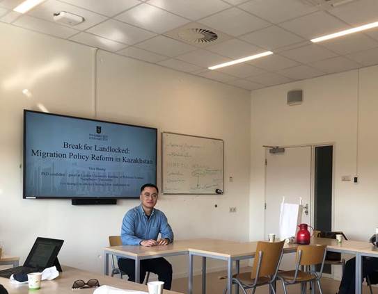 Yize Huang sitting by the desk in a room with chairs, desks, white walls, presentation slides on the screen on the wall